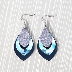 May include: A pair of silver dangle earrings with three layers of blue and silver patterned leather. The top layer is silver glitter, the middle layer is blue with white dots, and the bottom layer is dark blue.