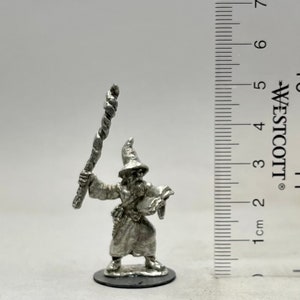 May include: A silver miniature figurine of a wizard holding a staff and a scroll. The wizard is wearing a pointed hat and a long robe. The figurine is standing on a round base.