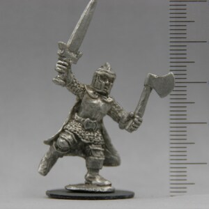 May include: A silver miniature figure of a warrior wearing armor and holding a sword and an axe. The figure is standing on a black base and is approximately 3 cm tall.