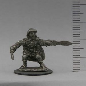 May include: A pewter miniature figure of a hooded person holding a dagger and a sword. The figure is standing on a round base.