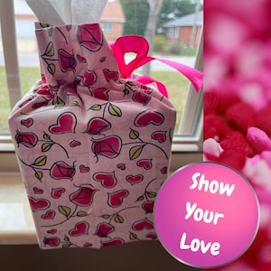 May include: A fabric tissue box cover featuring a light pink background with a repeating pattern of dark pink roses, green leaves, and small hearts. The cover is cinched at the top with a bright pink ribbon bow. A circular graphic displays the text Show Your Love.
