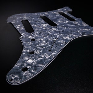 May include: A white pearlescent guitar pickguard with a black outline and multiple screw holes.