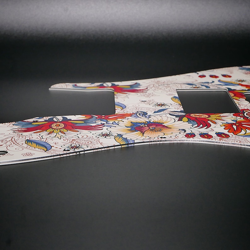 Puede incluir: Una placa de protecci&oacute;n de guitarra blanca con un patr&oacute;n floral en tonos rojos, azules y amarillos. La placa de protecci&oacute;n tiene tres recortes para los botones.