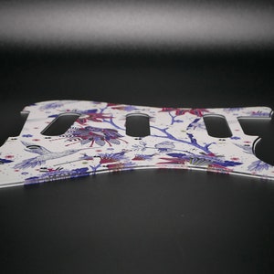 Pu&ograve; includere: Battuta per chitarra bianca con un motivo floreale che presenta colibr&igrave;, rami e fiori in tonalit&agrave; di viola, rosa e blu.