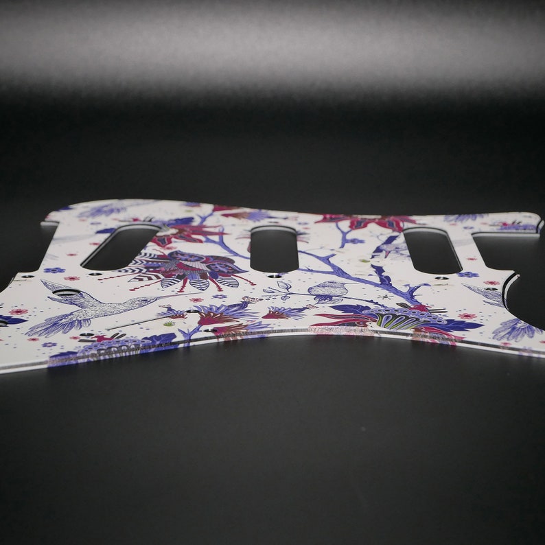 Pu&ograve; includere: Battuta per chitarra bianca con un motivo floreale che presenta colibr&igrave;, rami e fiori in tonalit&agrave; di viola, rosa e blu.