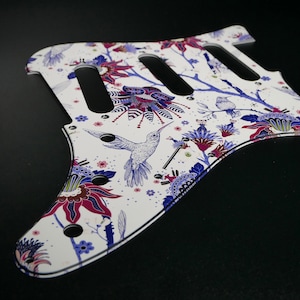 Pu&ograve; includere: Battuta per chitarra bianca con un design floreale e colibr&igrave;. La battuta presenta uno sfondo bianco con fiori blu, viola e rosa e colibr&igrave;.