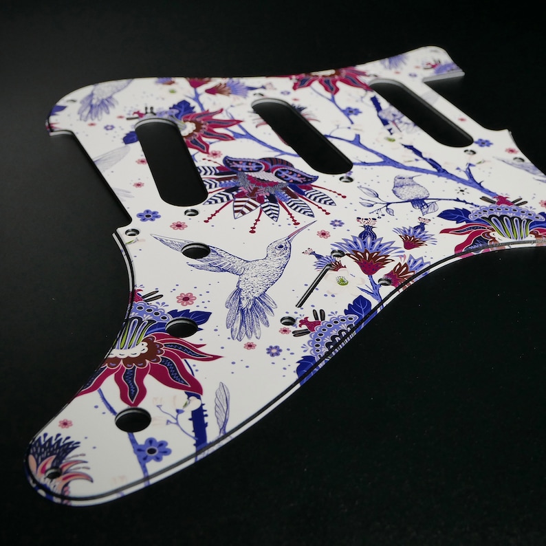 Pu&ograve; includere: Battuta per chitarra bianca con un design floreale e colibr&igrave;. La battuta presenta uno sfondo bianco con fiori blu, viola e rosa e colibr&igrave;.