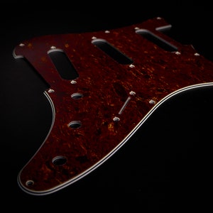 Peut inclure: Un pickguard de guitare marron en écaille de tortue avec une bordure blanche. Le pickguard comporte plusieurs trous pour les micros et les vis, ainsi que trois longues découpes rectangulaires. Le pickguard est fait d'un matériau à motifs.