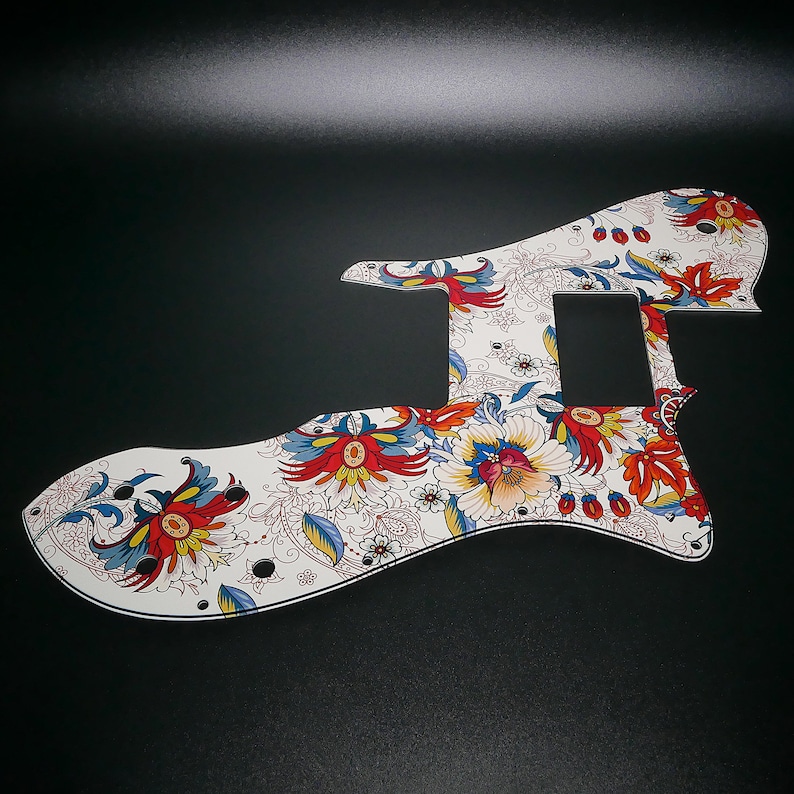 Puede incluir: Golpeador de guitarra blanco con un patr&oacute;n floral en rojo, azul y amarillo. El golpeador tiene un recorte para una pastilla y una perilla de control.
