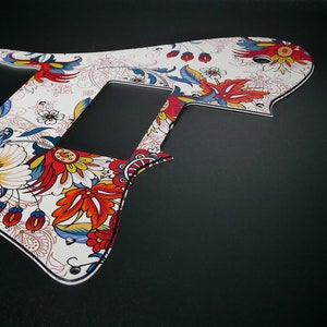Puede incluir: Un pickguard de guitarra blanco con un patr&oacute;n floral colorido. El patr&oacute;n presenta flores rojas, azules y amarillas sobre un fondo blanco.