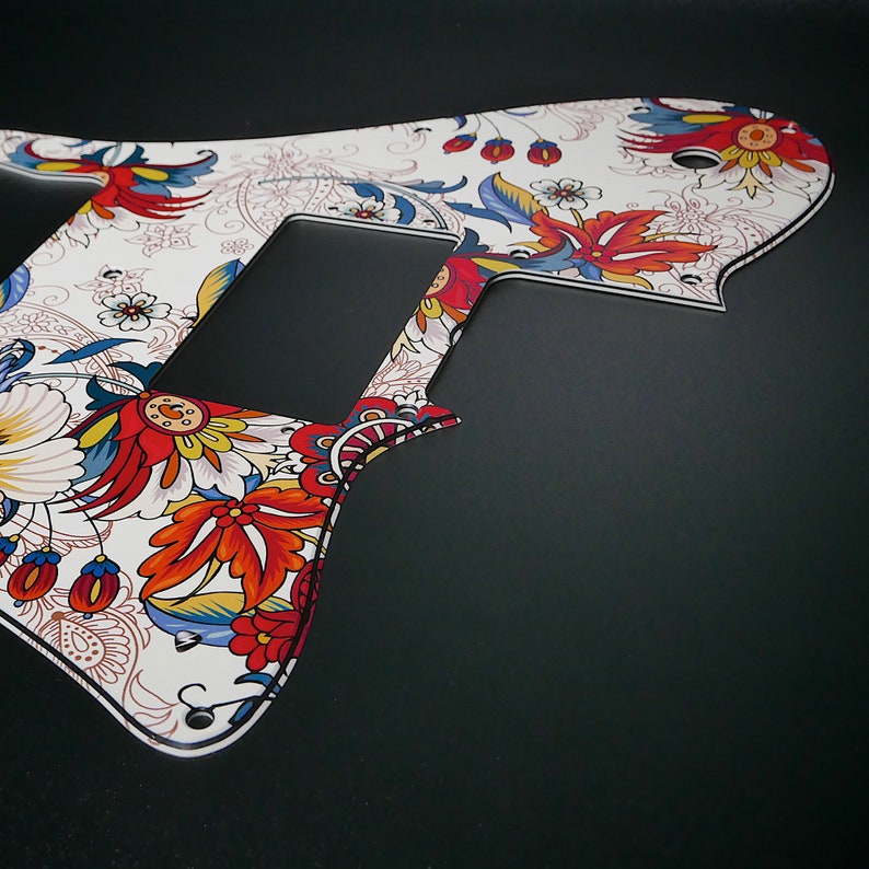 Puede incluir: Un pickguard de guitarra blanco con un patr&oacute;n floral colorido. El patr&oacute;n presenta flores rojas, azules y amarillas sobre un fondo blanco.