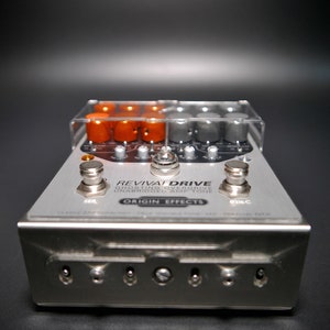 Könnte beinhalten: Ein silbernes Gitarreneffektpedal mit dem Text "REVIVAL DRIVE GHOSTING OVERDRIVE UNABRIDGED AMPTONE ORIGIN EFFECTS" auf der Vorderseite. Das Pedal hat eine transparente Plastikabdeckung mit orangefarbenen und silbernen Knöpfen.