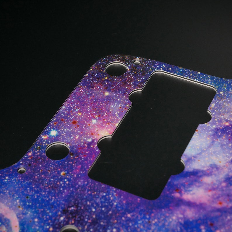 Op de afbeelding: Een paarse en blauwe gitaar pickguard met een galaxy print. De pickguard heeft uitsparingen voor de knoppen en pickups van de gitaar.