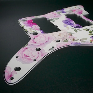 May include: White guitar pickguard with a floral pattern featuring pink and purple roses. The pickguard has several holes for mounting screws.