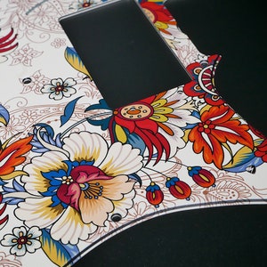 Puede incluir: Una placa de protecci&oacute;n de guitarra blanca con un patr&oacute;n floral colorido. El patr&oacute;n presenta flores rojas, amarillas, azules y verdes sobre un fondo blanco.