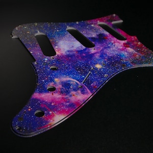 Peut inclure: Un pickguard de guitare avec un imprimé galaxie représentant une nébuleuse bleue, violette et rose avec des étoiles blanches.