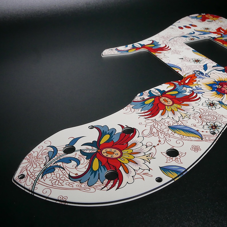 Puede incluir: Golpeador de guitarra blanco con un patr&oacute;n floral colorido. El dise&ntilde;o presenta flores rojas, azules, amarillas y blancas con detalles intrincados. El golpeador tiene varios agujeros negros para tornillos.
