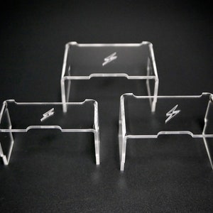 Op de afbeelding: Drie transparante acryl display stands met een bliksem logo. De stands zijn in verschillende maten en zijn in een rij op een zwart oppervlak gerangschikt.
