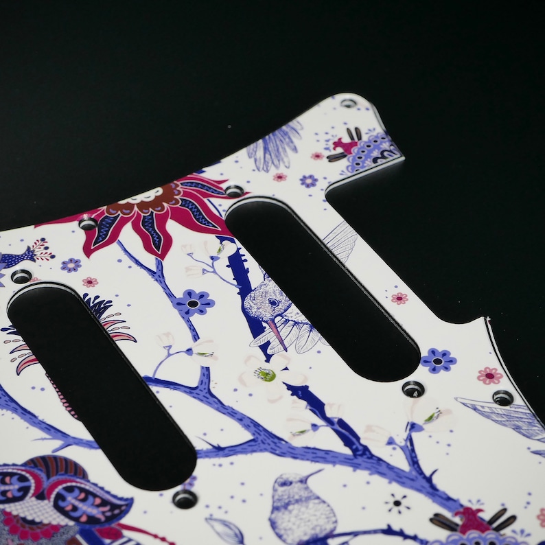 Pu&ograve; includere: Battuta di chitarra bianca con un design floreale con fiori blu e rosa, rami e uccelli. La battuta ha tre tagli per i pickup.