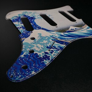Peut inclure: Un pickguard de guitare blanc et bleu avec un motif de grande vague, inspiré de la célèbre estampe japonaise "La Grande Vague de Kanagawa" d'Hokusai.