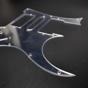 May include: A clear plastic guitar pickguard with multiple holes for mounting screws. The pickguard is shaped like a traditional electric guitar body.