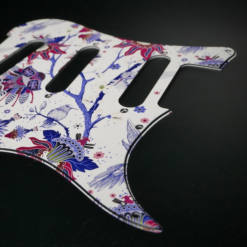 Pu&ograve; includere: Battuta di chitarra bianca con un motivo floreale in tonalit&agrave; di blu, rosa e viola. La battuta ha tre tagli per i pickup e uno sfondo bianco.