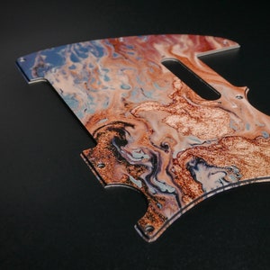 Peut inclure: Un pickguard de guitare avec un motif marbré aux nuances de bleu, brun et or. Le pickguard a une découpe pour un micro et un bouton de commande.