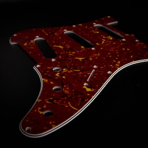 Peut inclure: Un pickguard de guitare électrique en écaille de tortue rouge et jaune. Le pickguard a trois découpes pour micros simple bobinage et une bordure blanche.