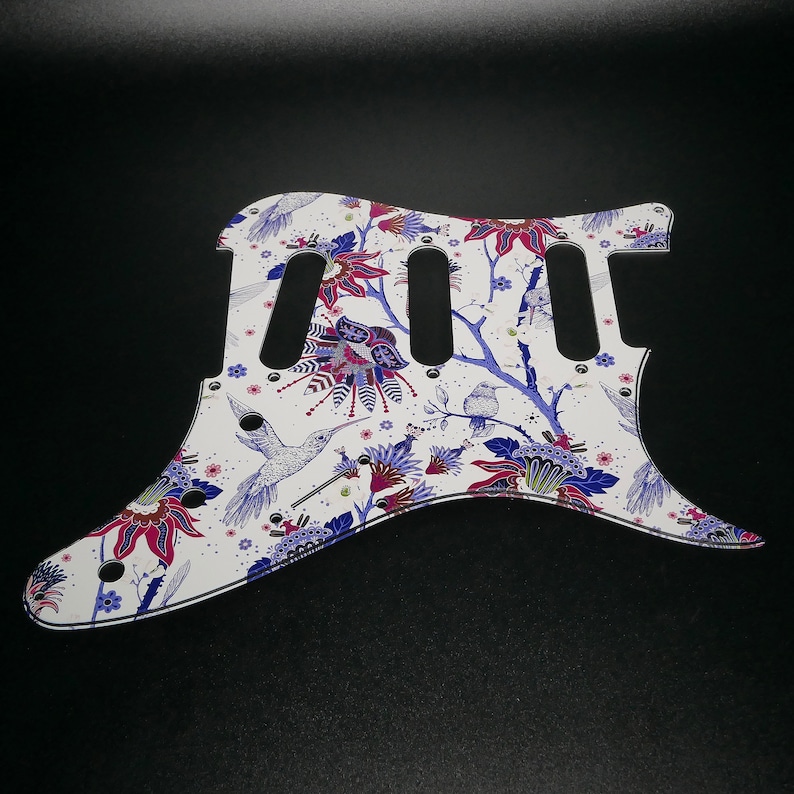 Pu&ograve; includere: Battuta per chitarra bianca con un design floreale e colibr&igrave; in tonalit&agrave; di blu, rosa e verde.