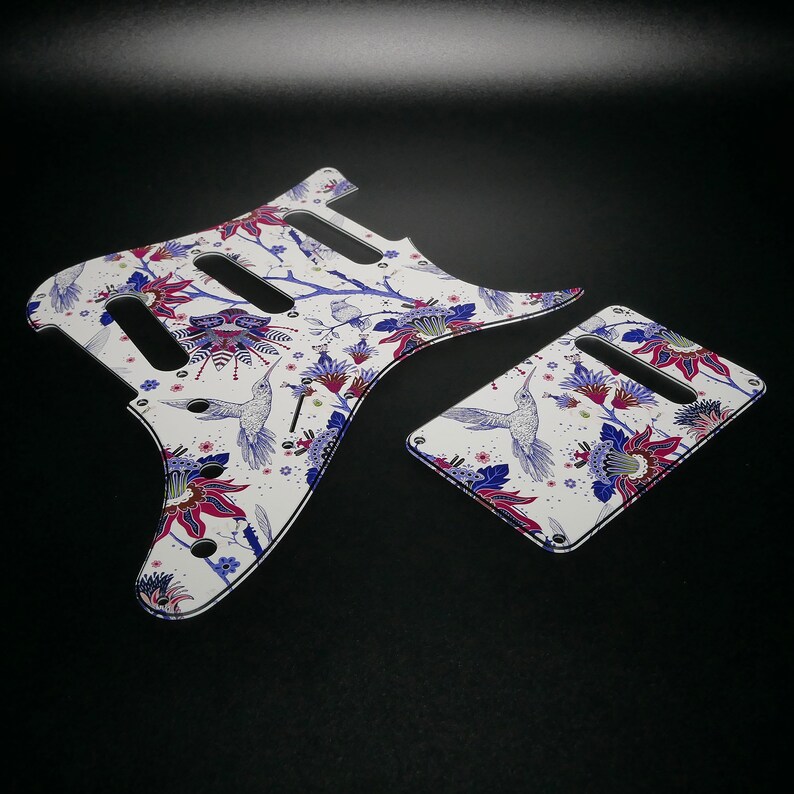 Pu&ograve; includere: Battuta per chitarra bianca con un motivo floreale che presenta colibr&igrave;, fiori e foglie in tonalit&agrave; di blu, viola e rosso.