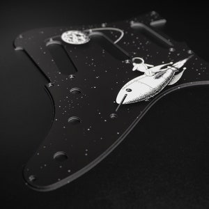 Peut inclure: Plaque de protection de guitare noire avec un astronaute blanc chevauchant une fusée dans l'espace. La plaque de protection a un motif de nuit étoilée avec une lune.
