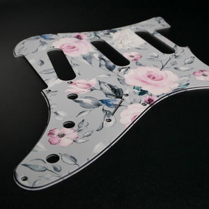 Peut inclure: Un pickguard de guitare gris avec un motif floral composé de roses roses et blanches, de feuilles vertes et d'accents bleus.