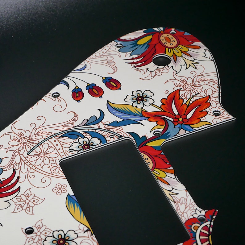 Puede incluir: Una placa de protecci&oacute;n de guitarra blanca con un patr&oacute;n floral colorido. El patr&oacute;n presenta flores y hojas rojas, azules, amarillas y blancas. La placa de protecci&oacute;n tiene dos recortes negros para las pastillas de la guitarra.