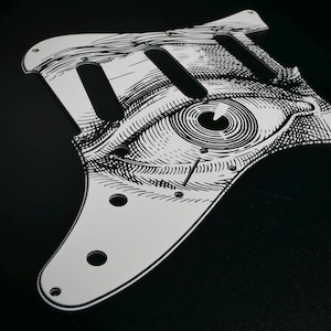Peut inclure: Un pickguard de guitare blanc avec une illustration noire et détaillée d'un œil. Le pickguard comporte trois découpes pour micros et des trous de vis. Le design rappelle une gravure.