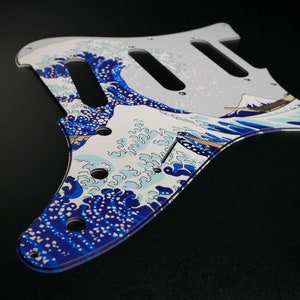 May include: A white and blue guitar pickguard with a design of a Japanese wave print. The pickguard is made of plastic and has a glossy finish.
