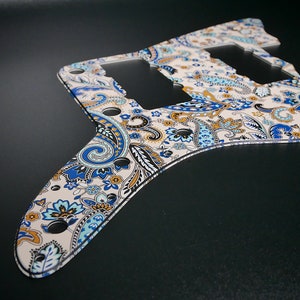 May include: A white guitar pickguard with a paisley pattern in blue, brown, and light blue. The pickguard has three cutouts for pickups and several small holes.