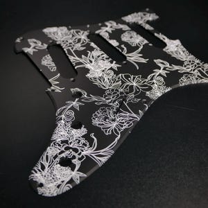 Peut inclure: Pickguard de guitare noir avec un motif floral blanc. Le pickguard a une forme unique avec trois découpes et plusieurs trous de vis. Le motif floral présente des contours détaillés de fleurs et de feuilles.