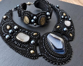 May include: A black beaded necklace and matching bracelet set featuring dark grey pearls, metallic gold accents, and large central druzy and agate stones. The intricate beadwork creates a textured, wide collar design with a secure metal clasp.