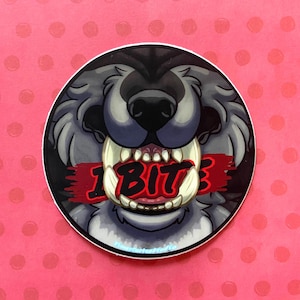 May include: A round sticker featuring a cartoon wolf's face with its mouth open, displaying large teeth. The wolf is gray and black, with the word "1 BIT" in red across its teeth. The sticker has a white border and is set against a pink background with small, darker pink polka dots.