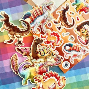 May include: A collection of die-cut stickers featuring various dinosaurs decorated with dessert elements like frosting, sprinkles, and fruit. The designs include a brown T-Rex, a white dinosaur with a cake-like back, and a rainbow-colored triceratops.