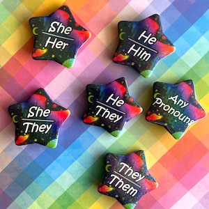 May include: Six star-shaped pins featuring a colorful galaxy print with planets and stars. Each pin displays white text indicating pronouns: She/Her, He/Him, She/They, He/They, Any Pronouns, and They/Them, separated by a horizontal white line.