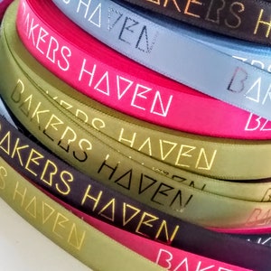 May include: A selection of colourful satin ribbons, including pink, blue, and green, are shown in a close-up shot. The ribbons are printed with the words "BAKERS HAVEN" in a gold metallic font. The ribbons are neatly coiled.