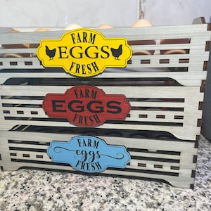 May include: A wooden egg crate with three levels, painted in light gray. Each level has a decorative sign that reads "FARM EGGS FRESH" in different colors: yellow, red, and blue. The crate is filled with brown eggs.