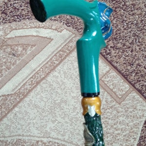 Puede incluir: Un bast&oacute;n de caminar verde azulado con un mango de madera tallada y un dise&ntilde;o verde y dorado. El mango tiene forma de cabeza de p&aacute;jaro con un pico azul.