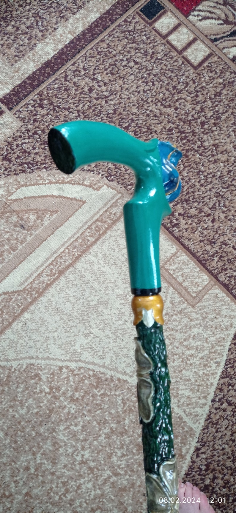 Puede incluir: Un bast&oacute;n de caminar verde azulado con un mango de madera tallada y un dise&ntilde;o verde y dorado. El mango tiene forma de cabeza de p&aacute;jaro con un pico azul.