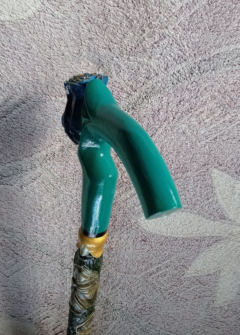 Puede incluir: Un bast&oacute;n de caminar verde azulado con un mango de madera tallado en forma de rosa. El mango est&aacute; pintado con detalles dorados y tiene una flor azul en la parte superior.