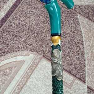 Puede incluir: Un bast&oacute;n de madera con un mango pintado de verde y azul con forma de rosa. El bast&oacute;n est&aacute; decorado con flores talladas y detalles dorados.