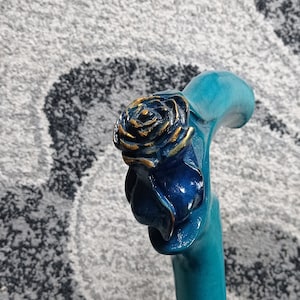 Puede incluir: Un bast&oacute;n de caminar azul turquesa con un dise&ntilde;o de rosa tallada. La rosa est&aacute; pintada de azul con detalles dorados.
