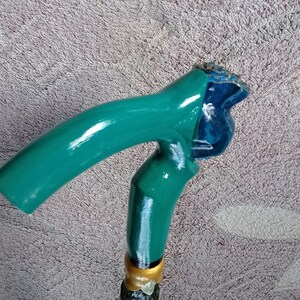 Puede incluir: Un bast&oacute;n de caminar verde azulado con un mango de madera tallado. El mango tiene forma de cabeza de p&aacute;jaro con un dise&ntilde;o azul y dorado.