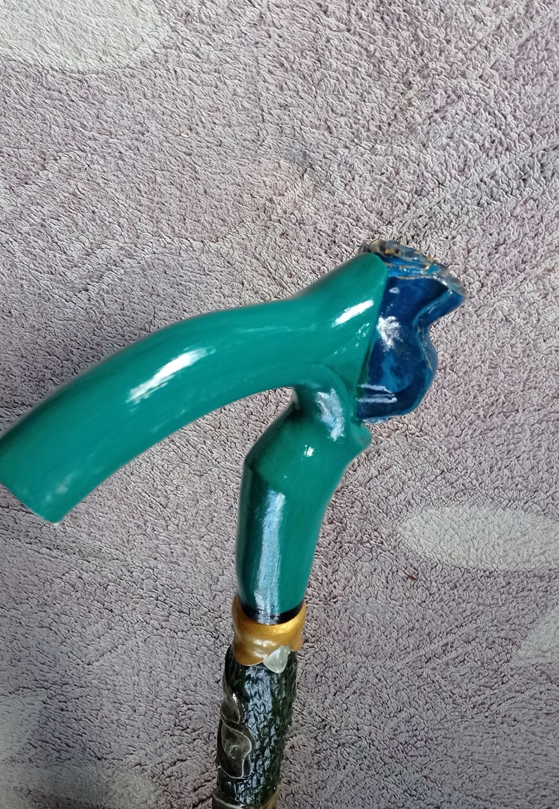 Puede incluir: Un bast&oacute;n de caminar verde azulado con un mango de madera tallado. El mango tiene forma de cabeza de p&aacute;jaro con un dise&ntilde;o azul y dorado.